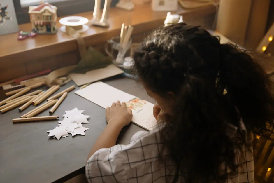 A young girl engaged in creative drawing of a Christmas card, surrounded by arts and crafts materials indoors.