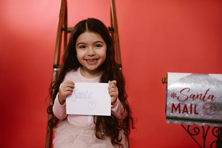 Adorable child with long hair smiling while holding a letter to Santa in a festive studio setting.