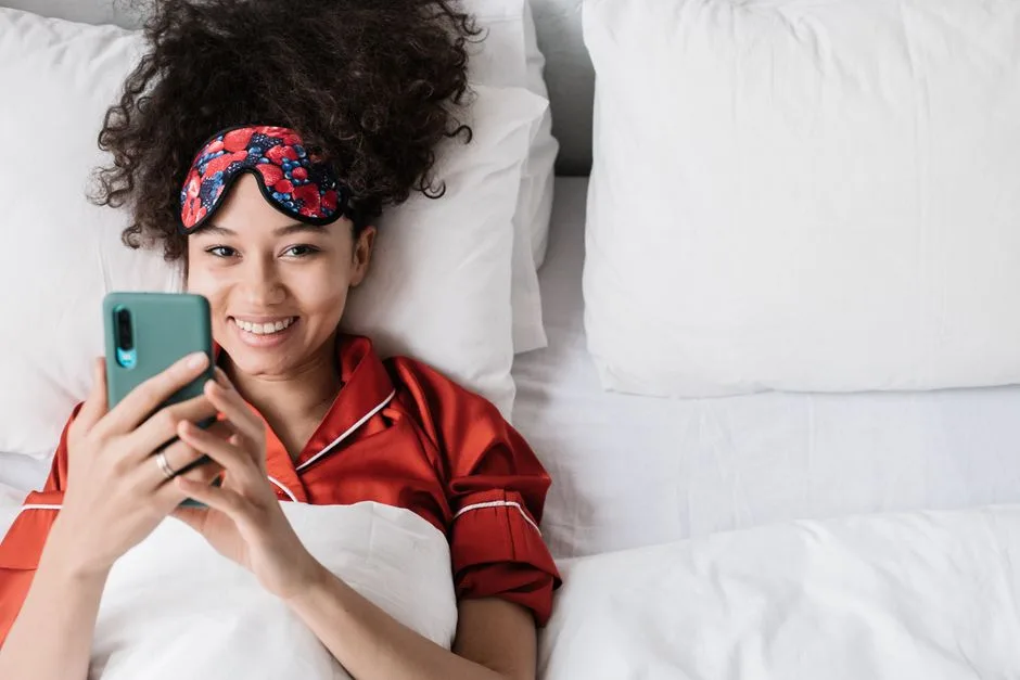 Happy woman with curly hair enjoying phone time in bed wearing sleep mask.