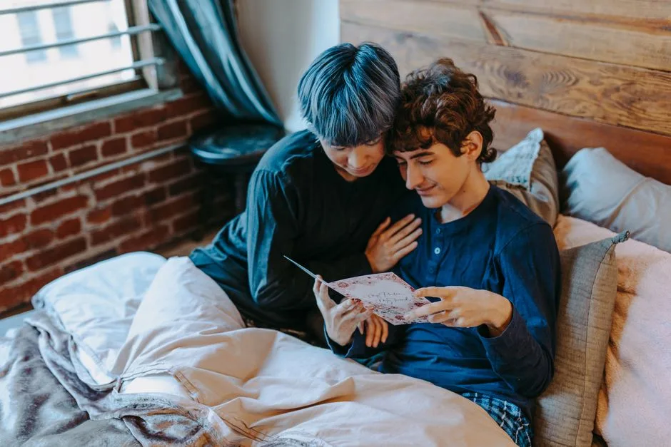 A loving gay couple reads a Valentine's Day card together in a cozy bedroom setting.