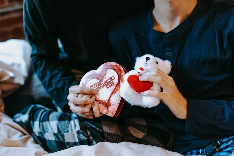A cozy indoor scene of adults exchanging Valentine's gifts, featuring a teddy bear and heart-shaped box.
