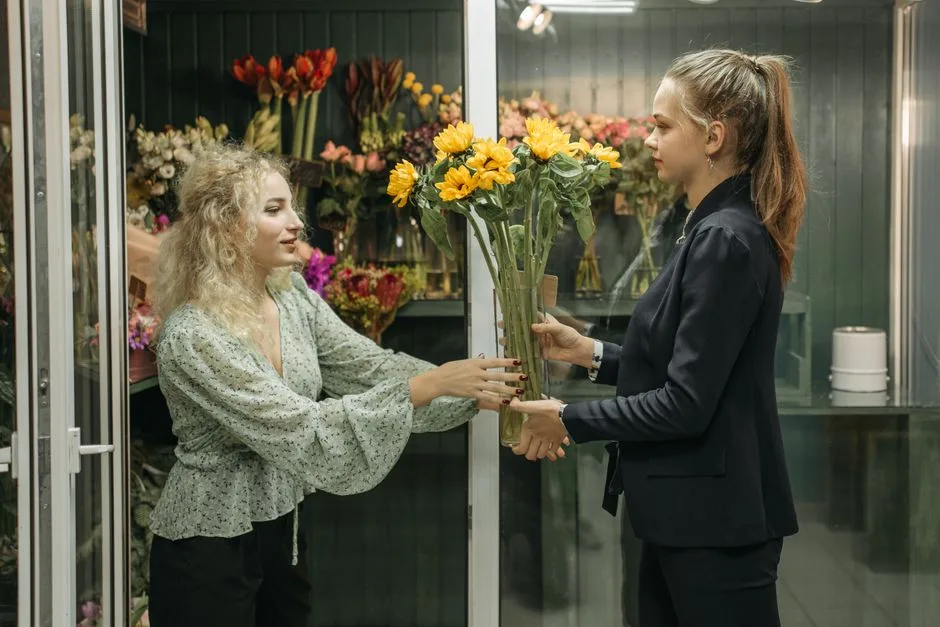 Two women standing, handing over sunflowers in a vibrant flower shop setting.