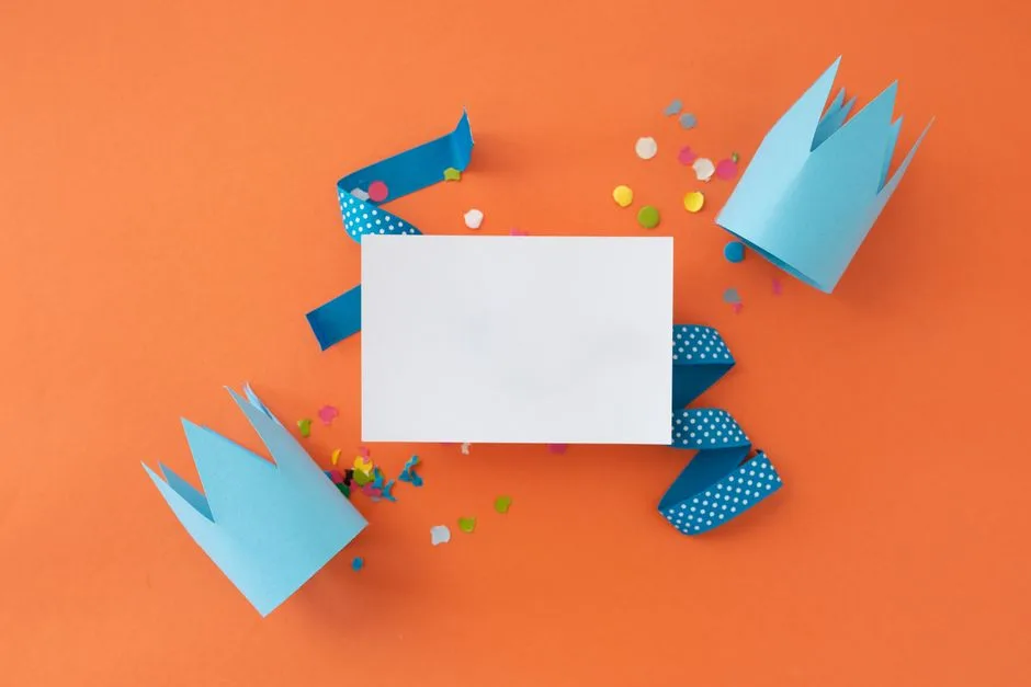 Flat lay of a blank card on orange with blue crowns, ribbons, and confetti, ideal for celebrations.