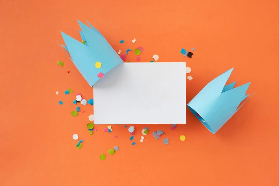 Colorful celebration scene with two paper crowns, confetti, and a blank card on vibrant orange.