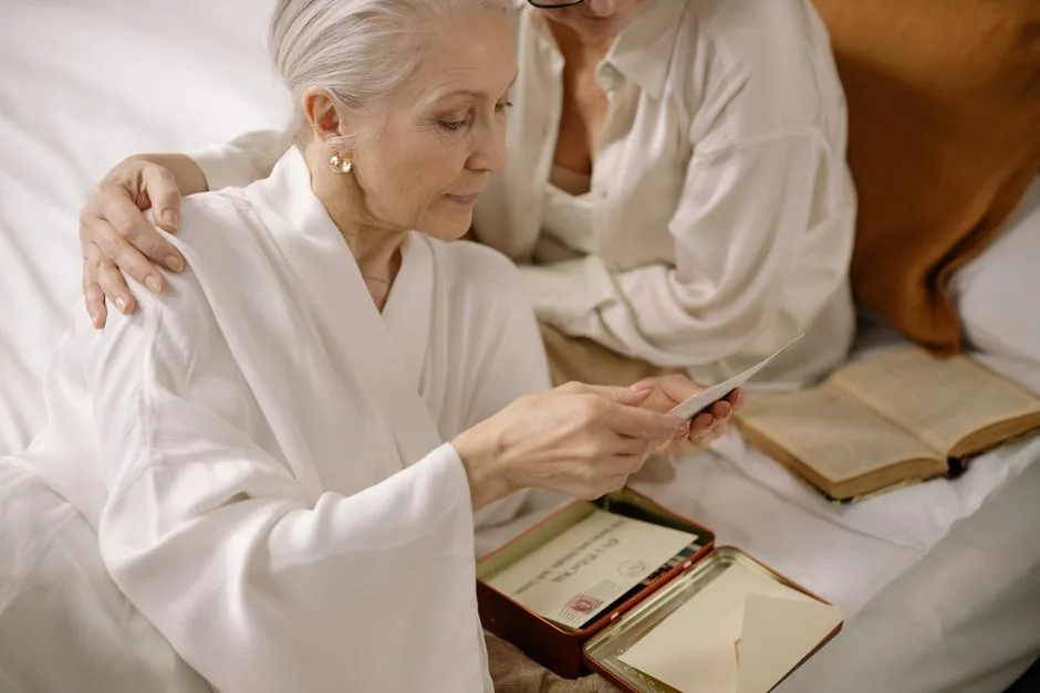 Elderly woman and companion reading old letters and photos on a bed, showcasing memories and togetherness.