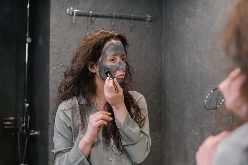 A woman applying a black facial mask while looking in the mirror in a contemporary bathroom.
