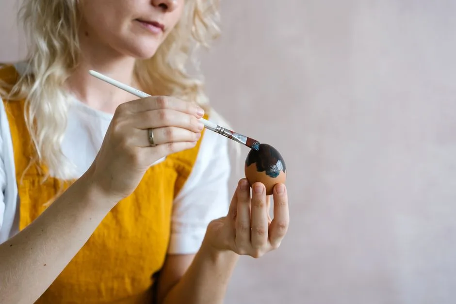 A woman in a yellow apron paints an Easter egg with black paint, capturing a festive DIY moment.