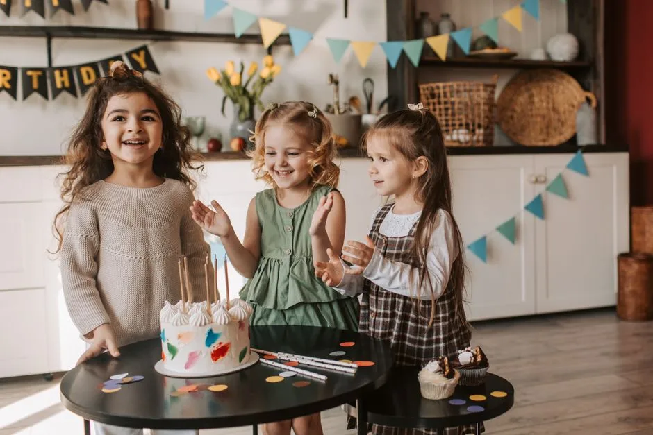 Three young girls enjoying a birthday party with a colorful cake and decorations indoors.