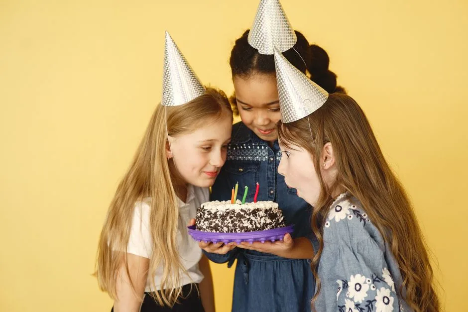 Three girls celebrate a birthday with hats and cake, against a yellow backdrop.