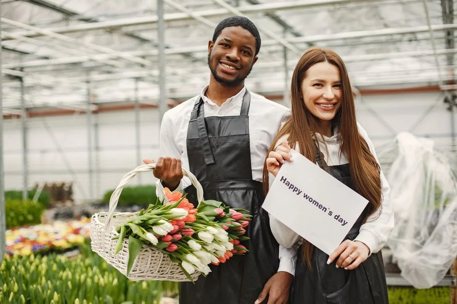 A smiling man and woman with flowers celebrate Women's Day in a greenhouse.