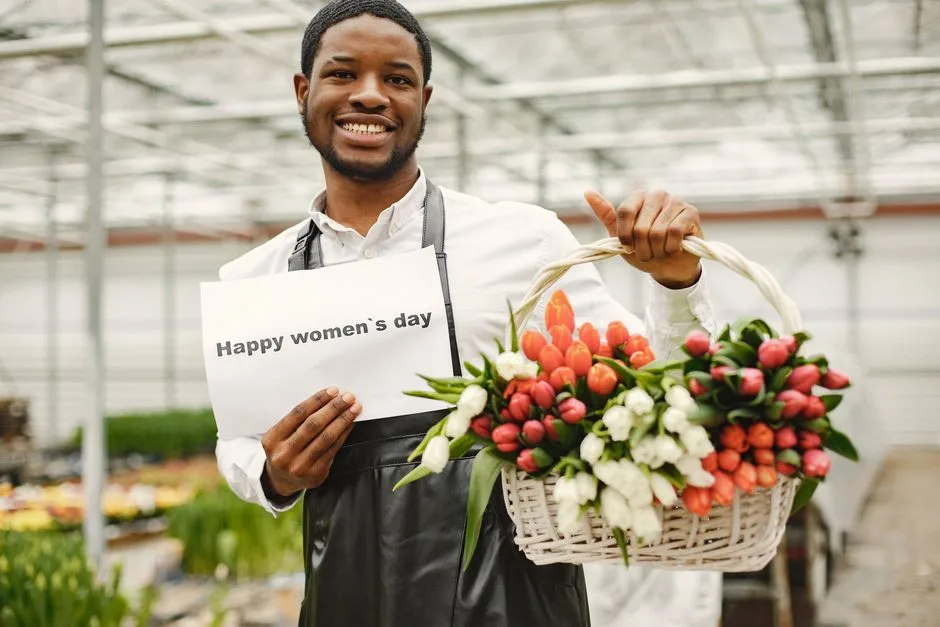 A cheerful gardener holding a basket of colorful tulips and a Happy Women's Day sign in a greenhouse.