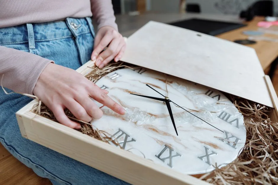 Hands gently inspecting a resin wall clock with Roman numerals in a wooden box, highlighting craftsmanship.