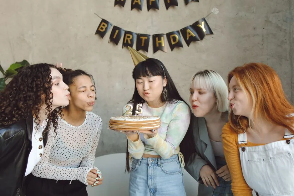 Group of diverse women celebrating a 25th birthday indoors with cake and decor.