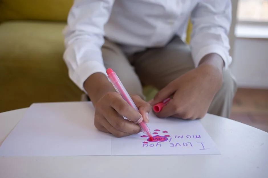 A close-up of a child writing 'I Love You Mom' on a card with a pink marker.