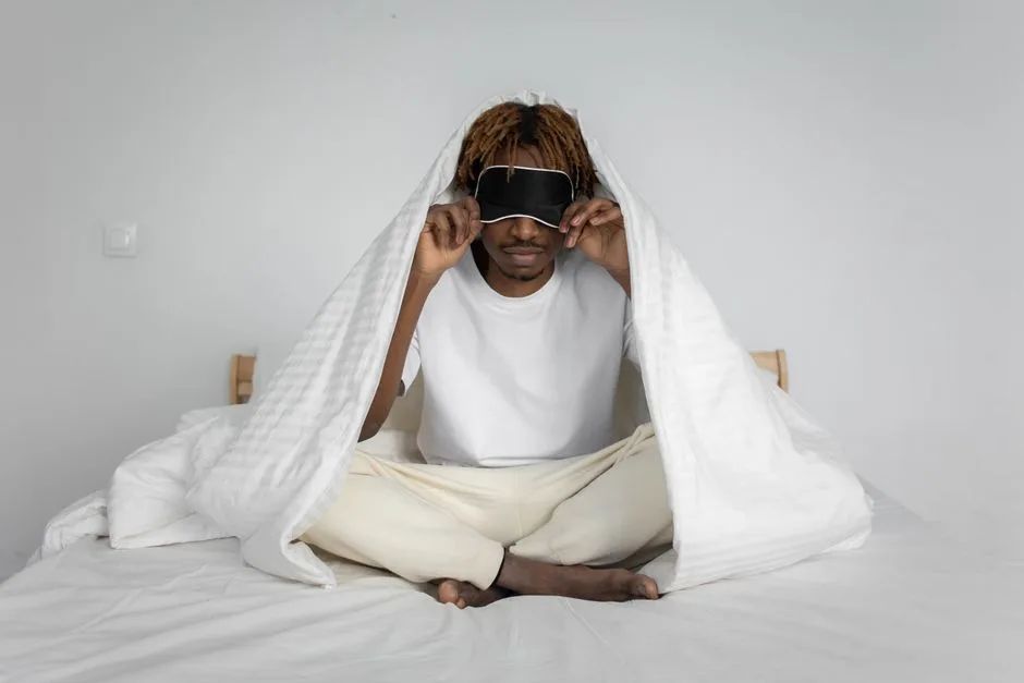 African American man sitting on bed with a sleep mask, wrapped in a comforter for relaxation.