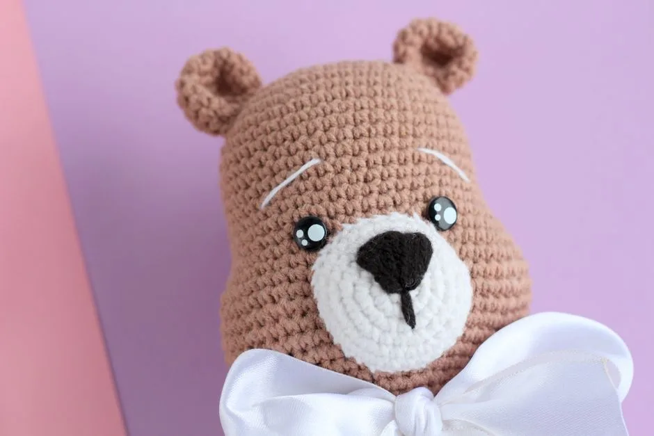 Close-up of a handmade crochet teddy bear with a white bow tie, set against pastel background.