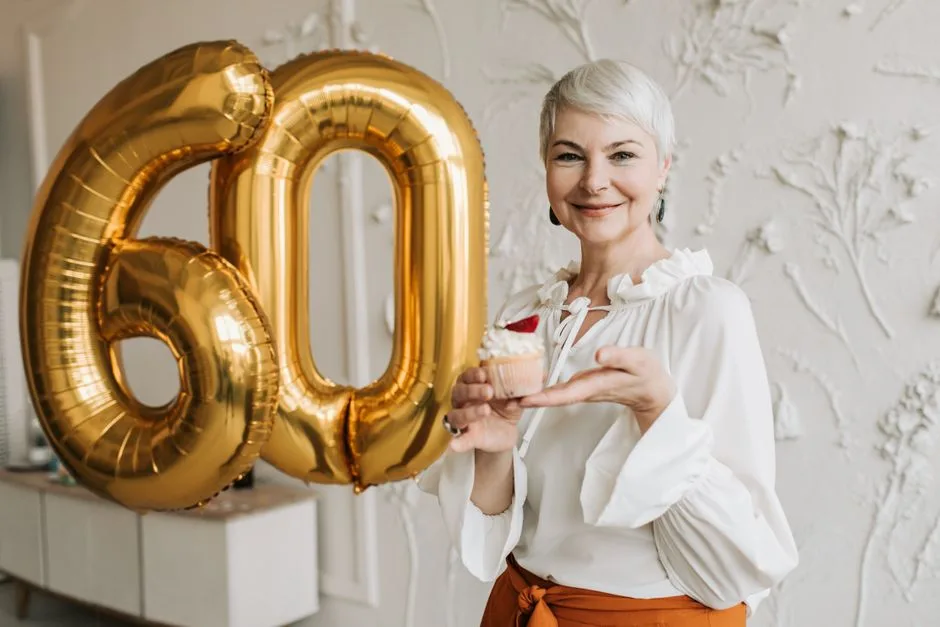 Senior woman with silver hair joyfully holding a cupcake next to 60 balloon indoors.