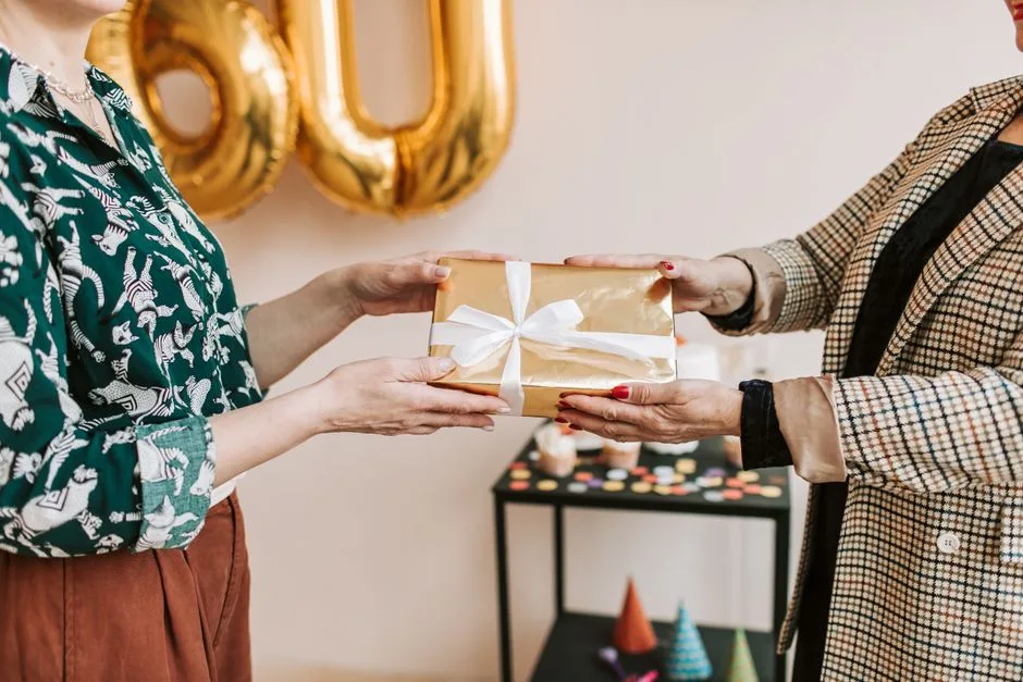 Two people exchanging a wrapped gift at a birthday celebration with decorations in the background.
