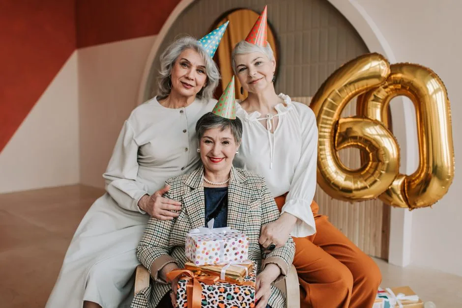 Three elegant women celebrate a 60th birthday with balloons and gifts indoors.