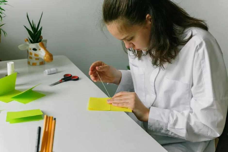 A young woman focuses intently on sewing a craft project at a brightly lit desk.
