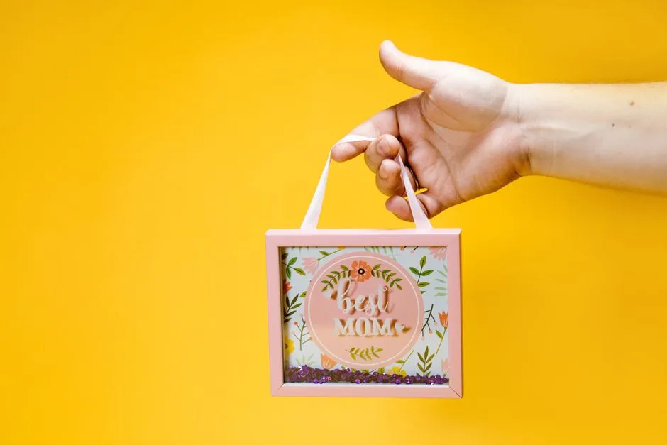 A hand holds a pink framed 'Best Mom' sign with floral design against a vibrant yellow background.