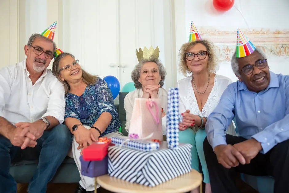 A joyful group of seniors wearing party hats and smiling while celebrating a birthday indoors.