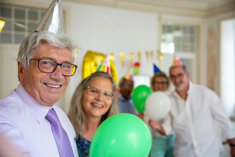 Seniors joyfully celebrating a birthday indoors with balloons and party hats.