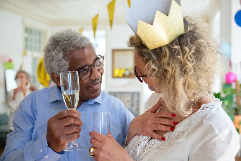 Elderly couple celebrating with champagne toast wearing party hats indoors.