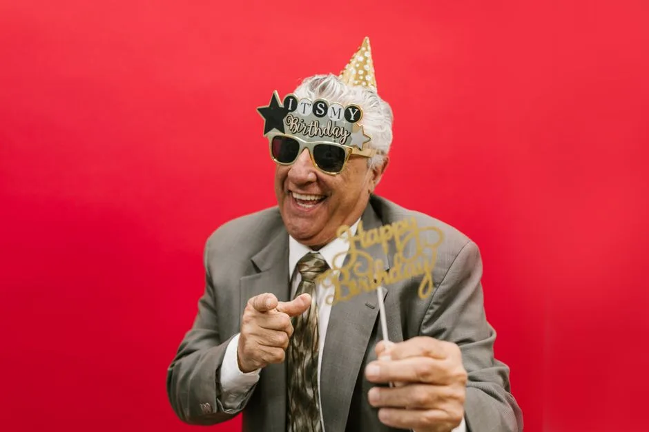 Senior man in festive attire enjoying his birthday with playful props against a vibrant red background.