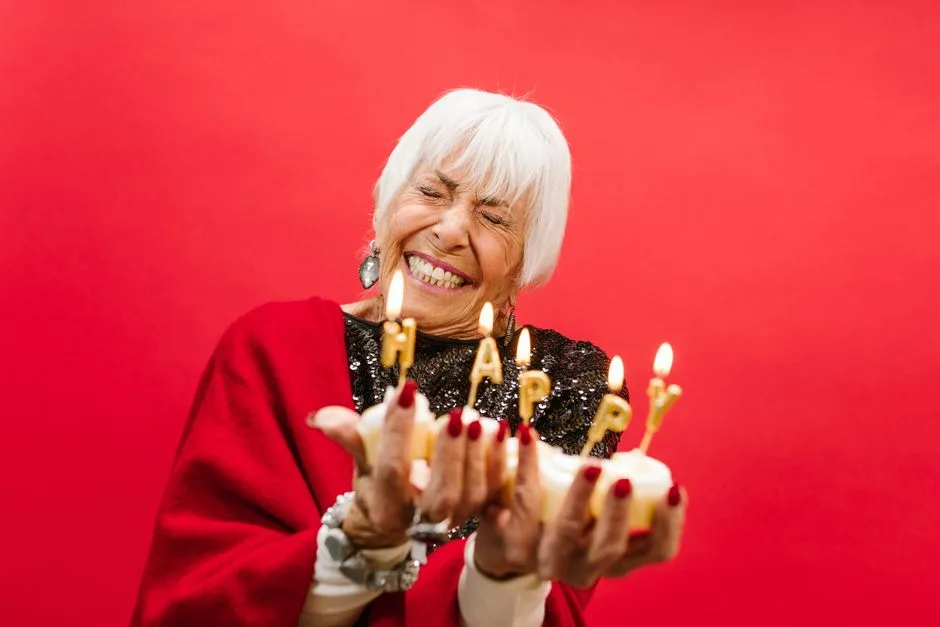 Cheerful elderly woman holding a birthday cake with lit candles spelling 'HAPPY' against a red background.