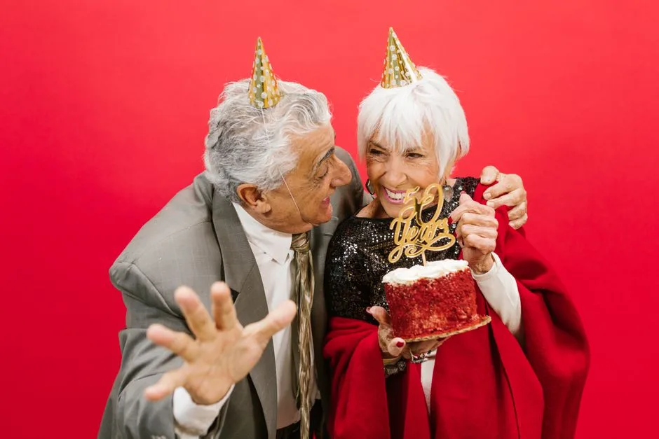 Elderly couple with party hats celebrating 50th anniversary with a cake and laughter.