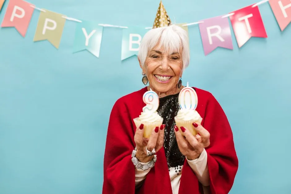 Smiling elderly woman holding cupcakes with candles on 90th birthday party.