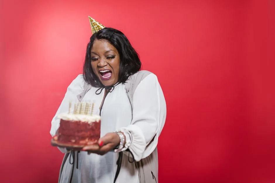 A joyful woman wearing a party hat holds a birthday cake against a vibrant red background.
