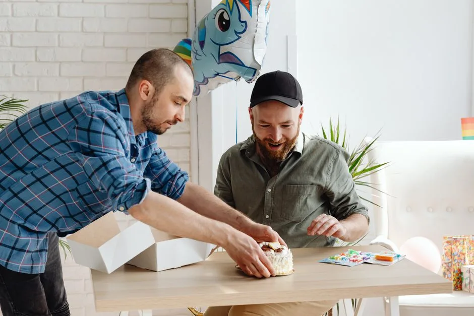Two men celebrating a birthday indoors with a cake, balloons, and gifts.