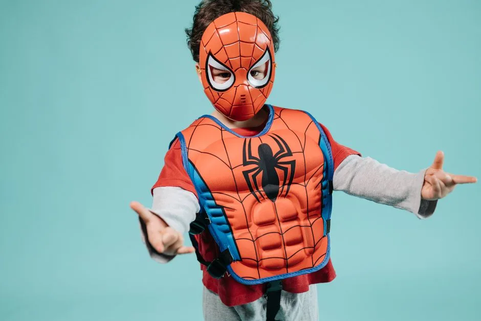 Child wearing a Spiderman costume and mask striking a playful superhero pose in a studio setting.