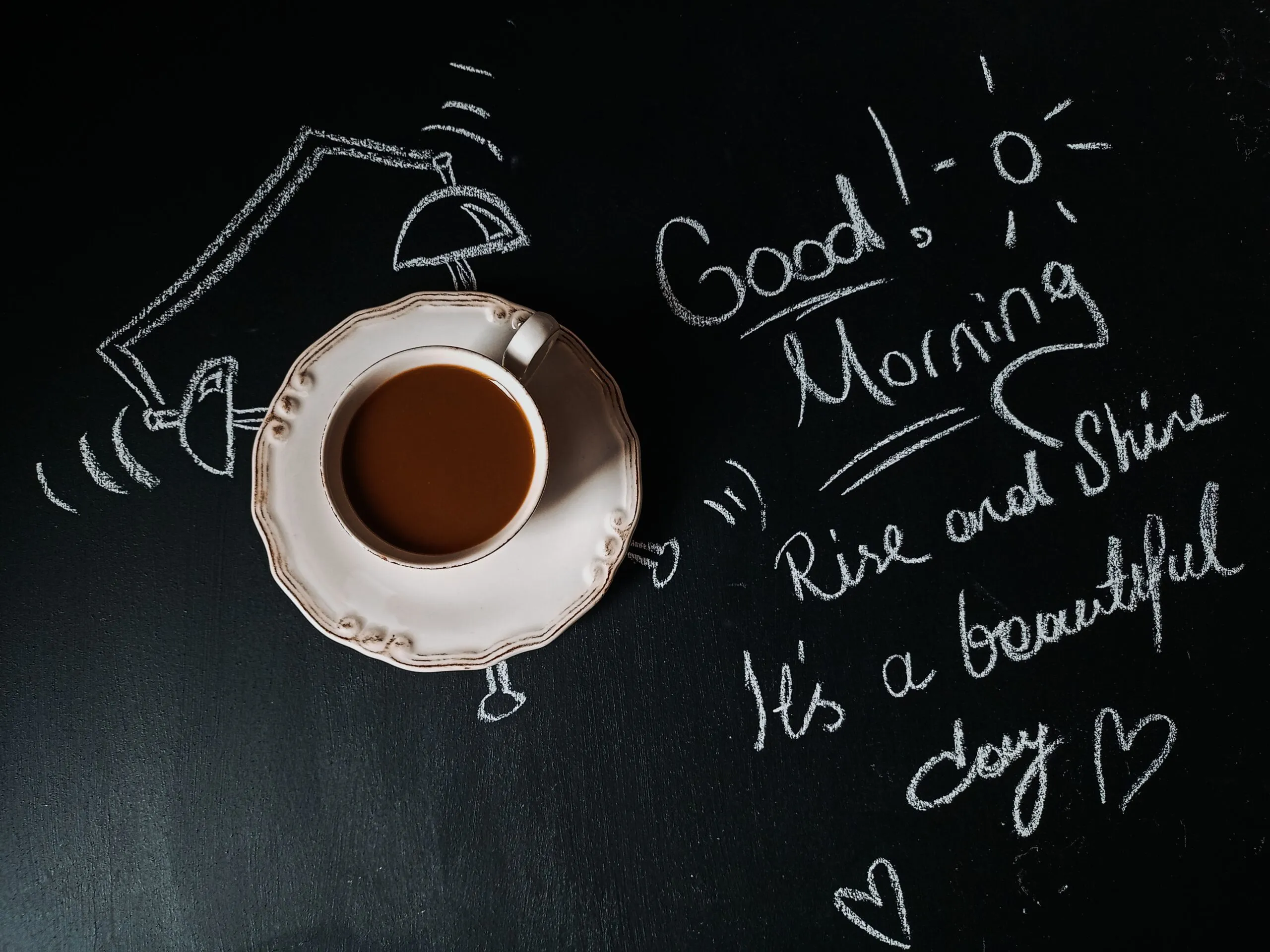 Cup of coffee on chalkboard with a cheerful good morning message and illustrations.