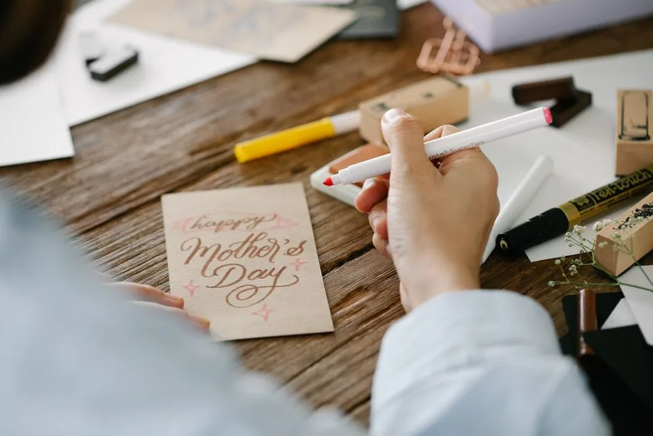 Hand lettering a personalized Mother's Day card with creative supplies on a wooden table.