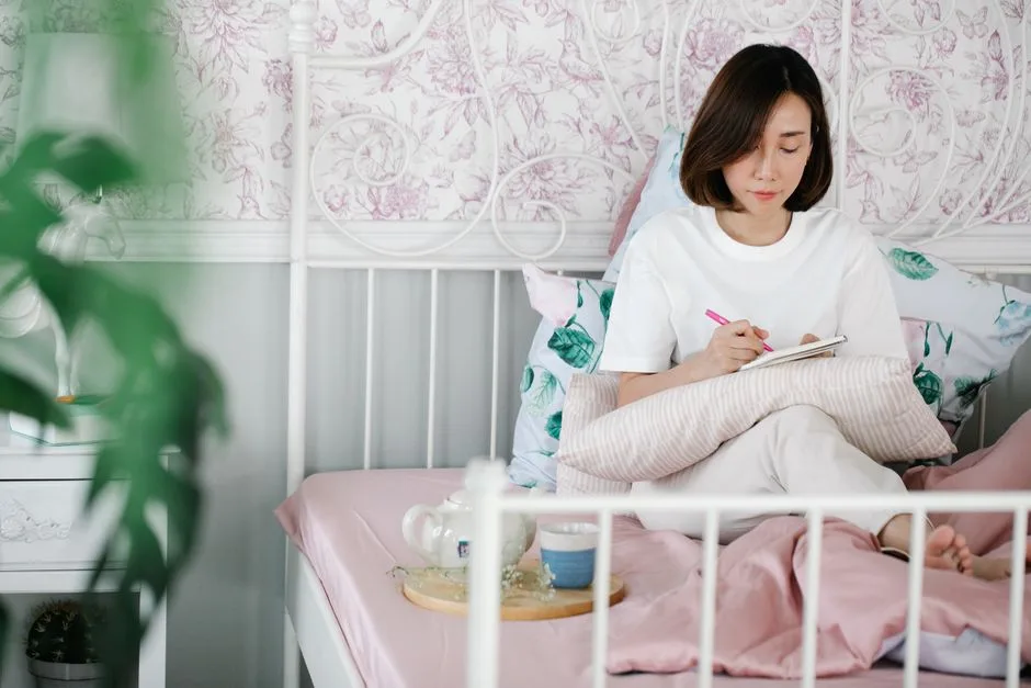 An Asian woman sitting on a bed, journaling in a serene and cozy bedroom environment.