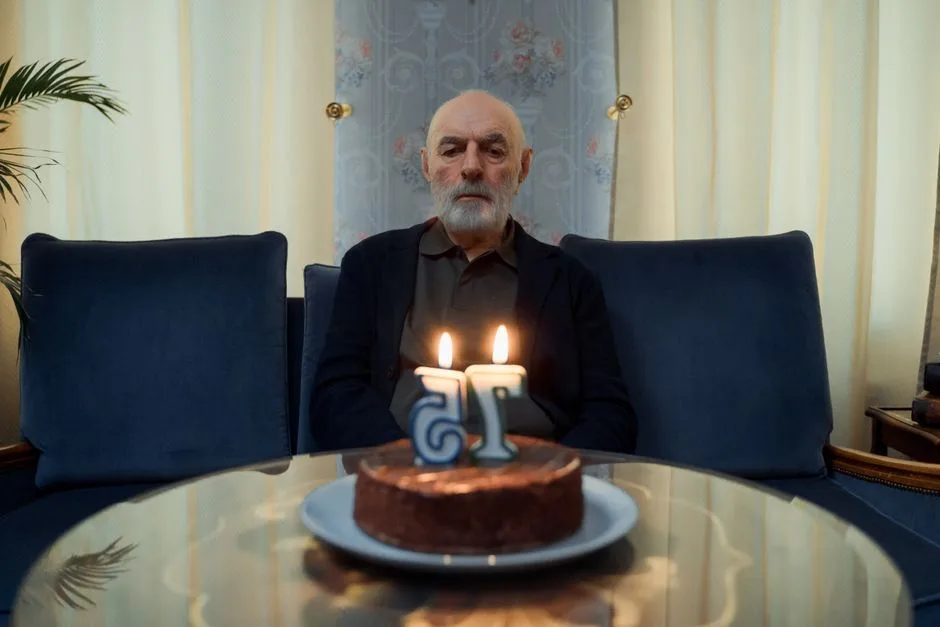 A senior man sitting in a living room with a birthday cake and candles on a glass table.