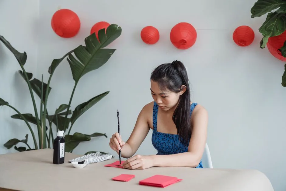 Woman in blue top creating art with red paper amidst lanterns and plants indoors.