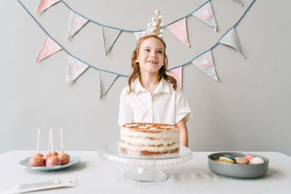 A smiling girl at a birthday party with a cake, wearing a party hat indoors.