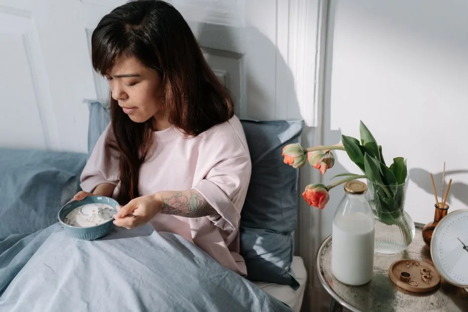 A woman sits in bed enjoying breakfast with milk and flowers nearby, bathed in morning light.