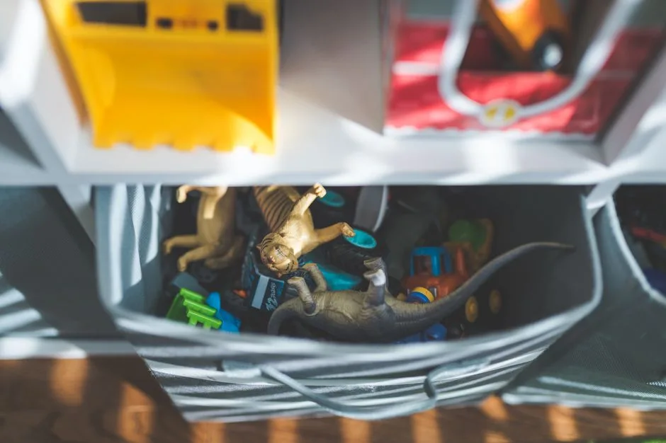 Plastic dinosaurs and cars in a storage box in a sunlit room.