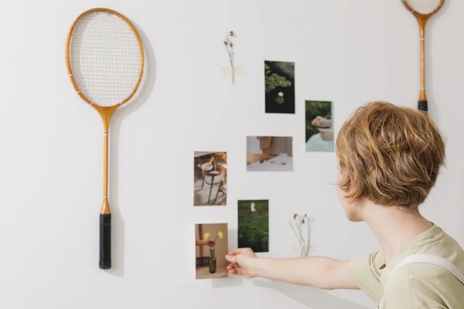 A woman arranges photos and tennis rackets on a wall, sparking creativity and inspiration.