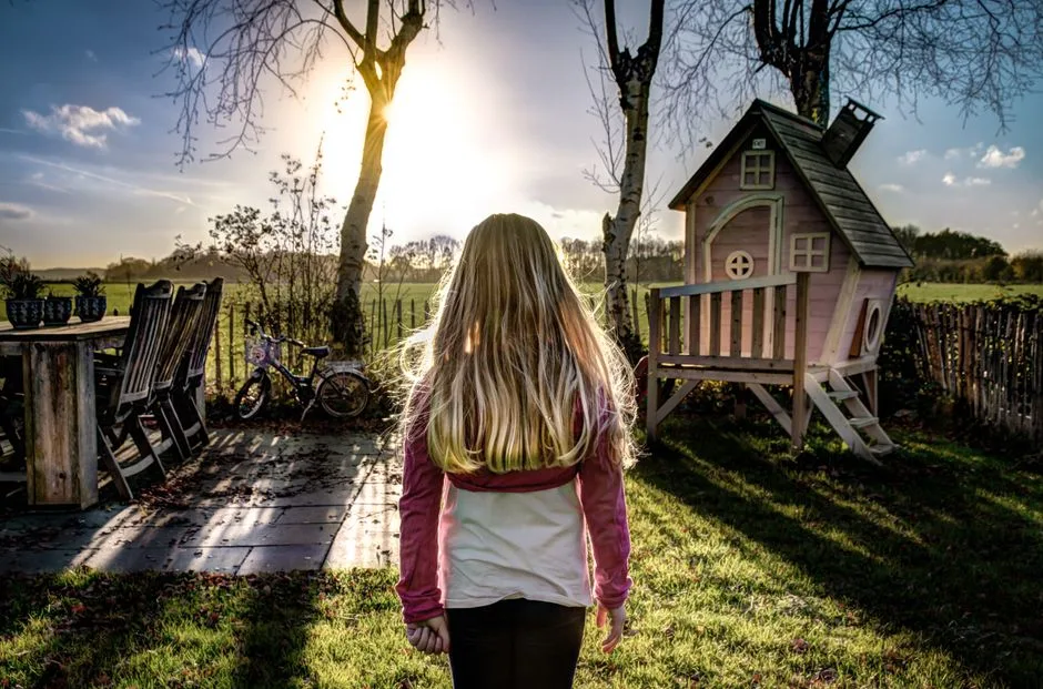 A child enjoying a sunny day in a backyard with a wooden playhouse and trees.