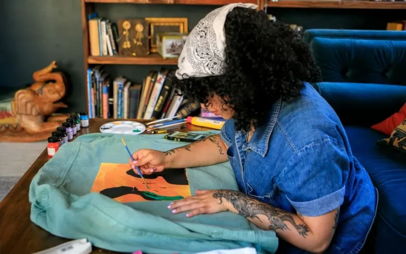A woman artist paints a vibrant design on fabric indoors, showcasing her skill and creativity.