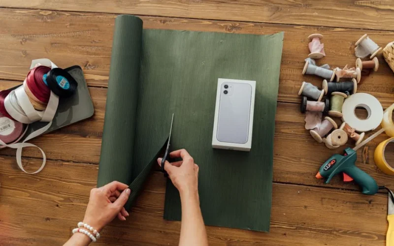 Hands wrapping a smartphone box with green paper on a wooden table, surrounded by ribbons and tools.