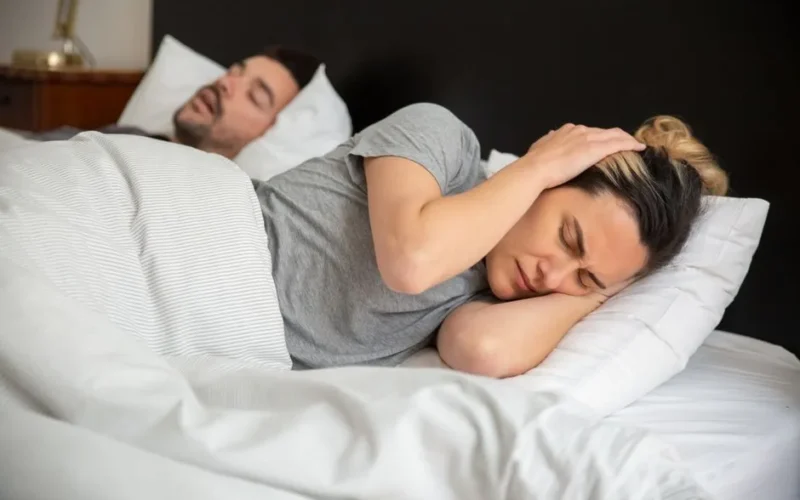 A woman looks frustrated in bed as her partner snores loudly, highlighting sleep disturbance issues.