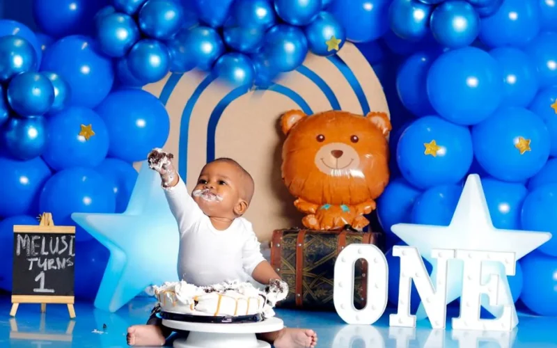 Adorable baby enjoying first birthday cake amidst blue balloon decorations.