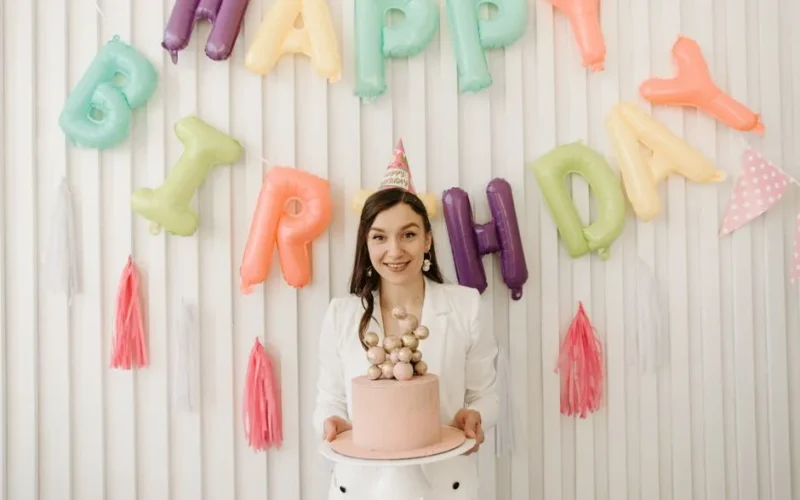 Woman celebrating birthday with colorful balloon decorations and a cake indoors.